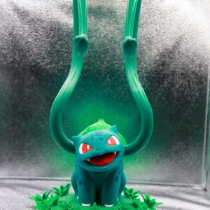 May include: A teal Bulbasaur figurine with red eyes and an open mouth, set on a green base with leafy details. Two long, curved green vines extend upwards from the figure's back. The background is a gradient of silver and green.