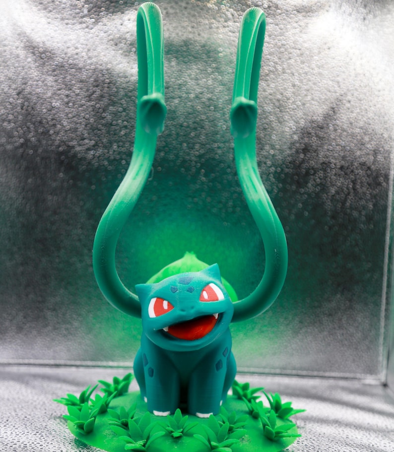 May include: A teal Bulbasaur figurine with red eyes and an open mouth, set on a green base with leafy details. Two long, curved green vines extend upwards from the figure's back. The background is a gradient of silver and green.