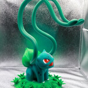 May include: A teal and green Bulbasaur figurine with red eyes and a large green bulb on its back. The figurine has two long, looping green vines extending upwards. It sits on a green base with grass details.