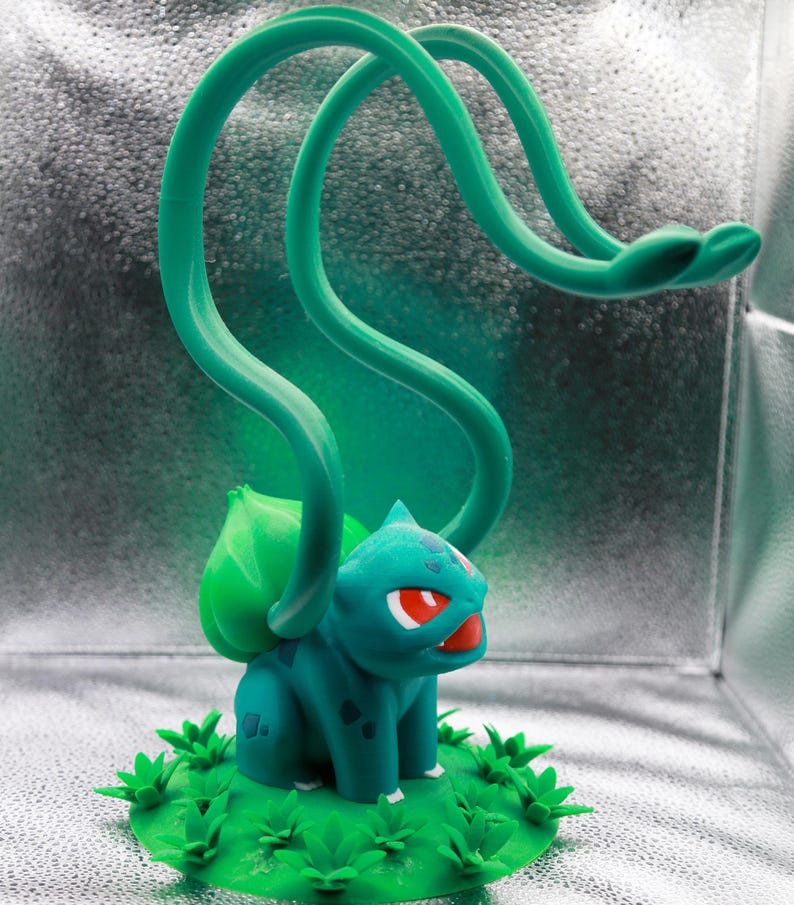May include: A teal and green Bulbasaur figurine with red eyes and a large green bulb on its back. The figurine has two long, looping green vines extending upwards. It sits on a green base with grass details.
