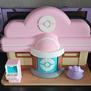 May include: A pink and white 3D-printed model of a Pokémon Center, featuring a blue and white Poké Ball design. Includes a small pink arcade machine and a purple Ditto figure. The model is set on a wooden base.