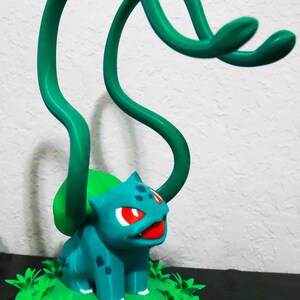 May include: A teal Bulbasaur figurine with red eyes and a red mouth, set on a green base with leafy details. Two green vines extend upwards from its back. The figurine is a collectible item.