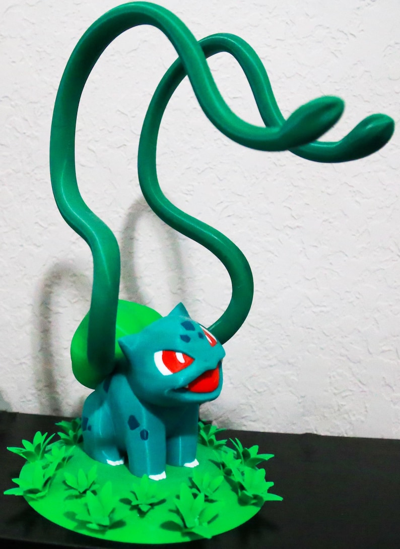 May include: A teal Bulbasaur figurine with red eyes and a red mouth, set on a green base with leafy details. Two green vines extend upwards from its back. The figurine is a collectible item.