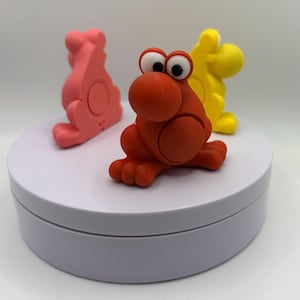 May include: Three colorful, cartoonish frog figurines in red, pink, and yellow. The red frog is in the foreground with large googly eyes. The other two frogs are in the background. The figurines are sitting on a white, circular platform.