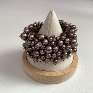 Op de afbeelding: Een clusterarmband met talrijke mauvekleurige imitatieparels van verschillende grootte. De armband heeft een vintage bronzen metalen ketting en is versierd met kleine, heldere kristallen. De armband wordt getoond op een witte, kegelvormige standaard.