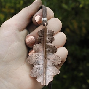 May include: A copper-colored leaf pendant with intricate veining details hangs from a brown cord. The pendant is held up by a silver-colored metal ring. The leaf design is a nature-inspired piece of jewelry.