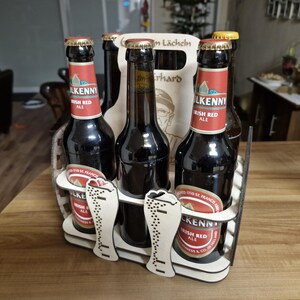 May include: A wooden beer caddy holding six bottles of Kilkenny Irish Red Ale. The bottles are dark with red and white labels. The caddy has a decorative cutout design with the words "Zum L&auml;cheln" and a portrait.