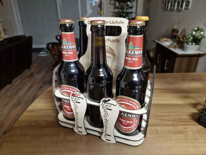 May include: A wooden beer caddy holding six bottles of Kilkenny Irish Red Ale. The bottles are dark with red and white labels. The caddy has a decorative cutout design with the words "Zum L&auml;cheln" and a portrait.