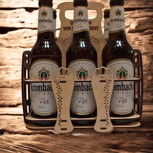 May include: A wooden beer carrier holding six bottles of Krombacher Pils beer. The bottles are brown with gold caps and white labels. Two wooden beer glass cutouts are in front of the bottles. The carrier has the text "D WIR N DIENT".