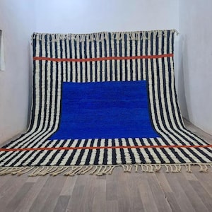 May include: A large rectangular area rug featuring a bold pattern of vertical black and white stripes, a prominent solid blue rectangular center, and two horizontal orange bands. The rug is finished with decorative fringe along the top and bottom edges.