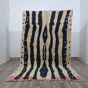 May include: A rectangular area rug featuring a cream-colored base with thick, vertical black stripes and a central diamond-shaped motif. The rug includes small black square accents along the edges and a horizontal pink band at the bottom fringe.