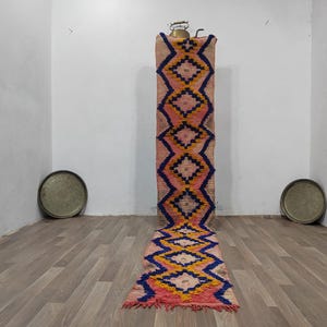 May include: A long, rectangular rug with a geometric diamond pattern in pink, blue, and yellow. The rug is displayed vertically, showcasing its design and texture. The rug is made of woven material.