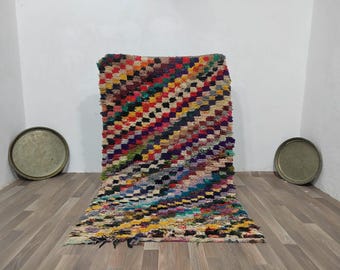 May include: A colorful, hand-woven rug with a geometric pattern. The rug features a variety of colors, including red, yellow, blue, and green. The rug is rectangular and has a textured surface. Two round metal trays are on the sides.