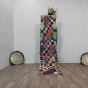 May include: A colorful, rectangular rug with a checkered pattern in shades of pink, blue, green, and brown. A small brass teapot sits atop the rug. Two round, metal trays are on the floor on either side of the rug.