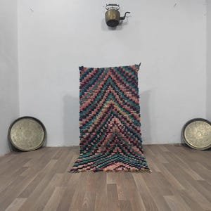 Op de afbeelding: Een kleurrijk, handgemaakt vloerkleed met een geometrisch chevronpatroon in roze, teal en zwart. Het vloerkleed wordt gepresenteerd tegen een witte muur, met twee decoratieve metalen trays op de vloer.