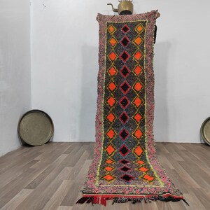 May include: A long, rectangular rug with a geometric diamond pattern in orange, black, and yellow on a gray background. The rug has a pink and green border with fringe at the bottom. The rug is displayed vertically.