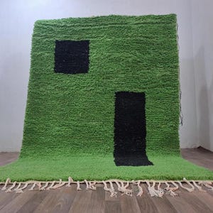 May include: A rectangular area rug with a thick, shaggy green pile featuring two distinct black geometric shapes: a square in the upper left and a vertical rectangle in the lower right. The rug is finished with off-white knotted fringe tassels.