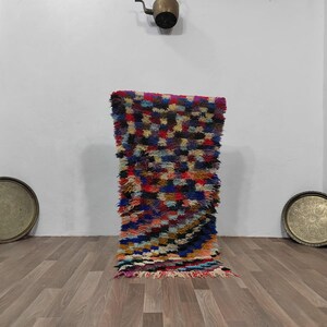 May include: A colorful, rectangular rug with a shaggy texture. The rug features a patchwork design with various colors, including red, blue, yellow, and black. Two brass trays are on the floor, and a brass kettle hangs on the wall.
