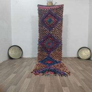 Può includere: Un tappeto rettangolare fatto a mano con un motivo a rombi nei toni del marrone, blu, rosso e verde. Il tappeto ha un aspetto strutturato ed è esposto verticalmente. Due vassoi rotondi in metallo sono sul pavimento.