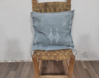 Puede incluir: Un cojín cuadrado azul claro con bordados geométricos blancos descansa sobre una silla tejida. El cojín presenta un diseño central y detalles en las esquinas. La silla tiene un marco de madera y un asiento y respaldo tejidos.