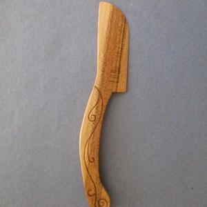 May include: A wooden butter knife with a curved handle and a decorative etched vine design. The knife is made of light brown wood and has a flat, angled blade. The handle curves gently, ending in a rounded tip.