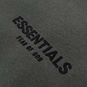 Puede incluir: Primer plano de una tela gris oscuro con la palabra "ESSENTIALS" y la frase "FEAR OF GOD" bordadas en negro. El texto est&aacute; en negrita, en una fuente sans-serif.
