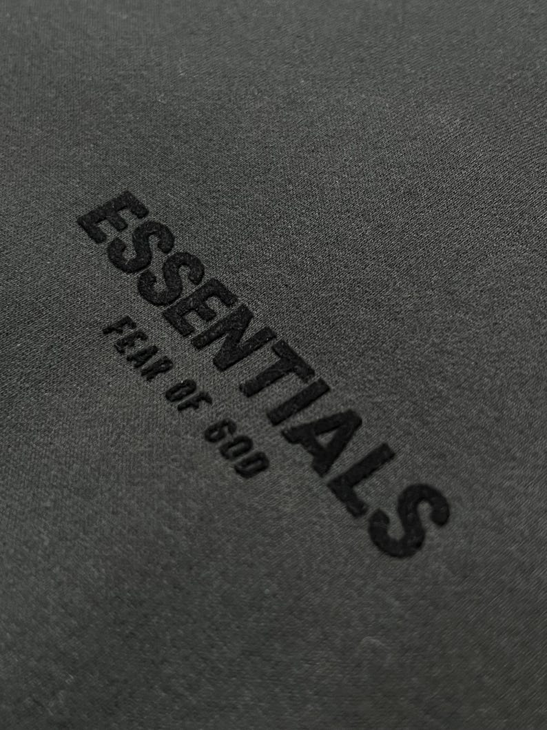 Puede incluir: Primer plano de una tela gris oscuro con la palabra "ESSENTIALS" y la frase "FEAR OF GOD" bordadas en negro. El texto est&aacute; en negrita, en una fuente sans-serif.