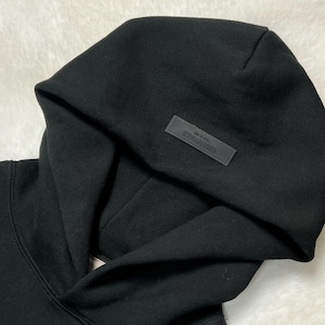 Puede incluir: Sudadera con capucha negra con una capucha grande. Un peque&ntilde;o parche rectangular con la palabra "ESSENTIALS" est&aacute; cosido en la capucha. La sudadera con capucha est&aacute; hecha de un material suave y grueso.