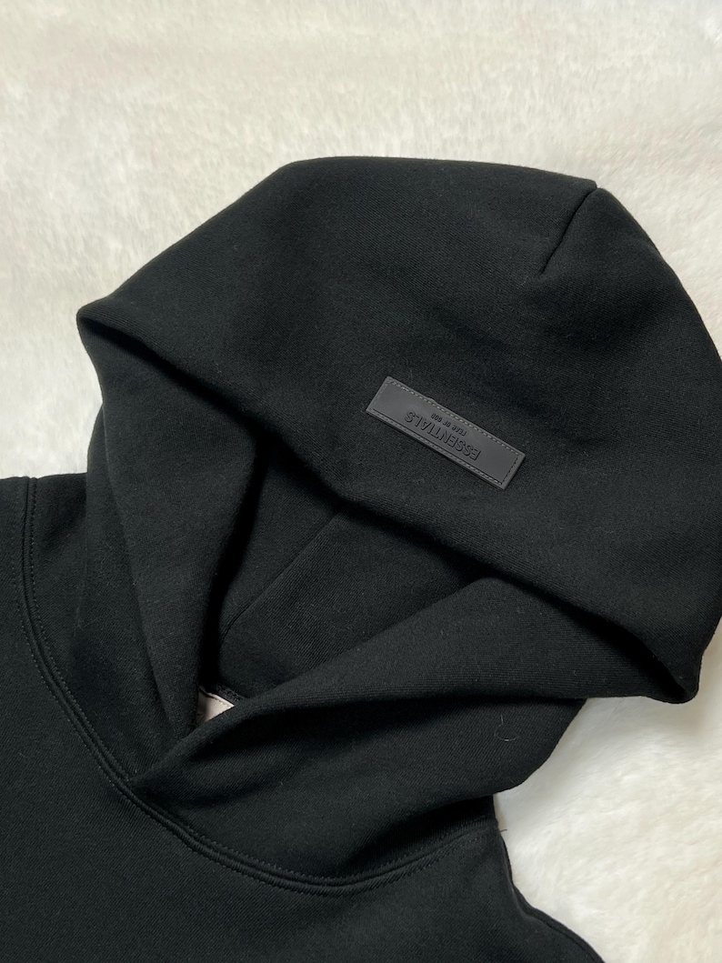 Puede incluir: Sudadera con capucha negra con una capucha grande. Un peque&ntilde;o parche rectangular con la palabra "ESSENTIALS" est&aacute; cosido en la capucha. La sudadera con capucha est&aacute; hecha de un material suave y grueso.