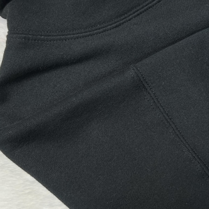 Puede incluir: Primer plano de una sudadera con capucha negra. La tela es de color negro liso con textura visible y detalles de costura. La imagen destaca la capucha y la zona de los hombros de la prenda.