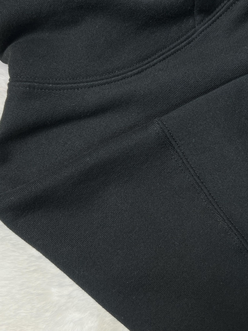 Puede incluir: Primer plano de una sudadera con capucha negra. La tela es de color negro liso con textura visible y detalles de costura. La imagen destaca la capucha y la zona de los hombros de la prenda.