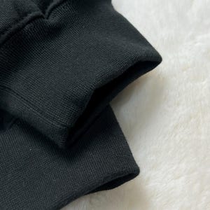 Puede incluir: Primer plano de un pu&ntilde;o de sudadera negro, que muestra la tela de punto acanalado y la costura donde se une a la manga. El pu&ntilde;o est&aacute; doblado, revelando el interior de la tela. El fondo es una superficie suave y blanca.