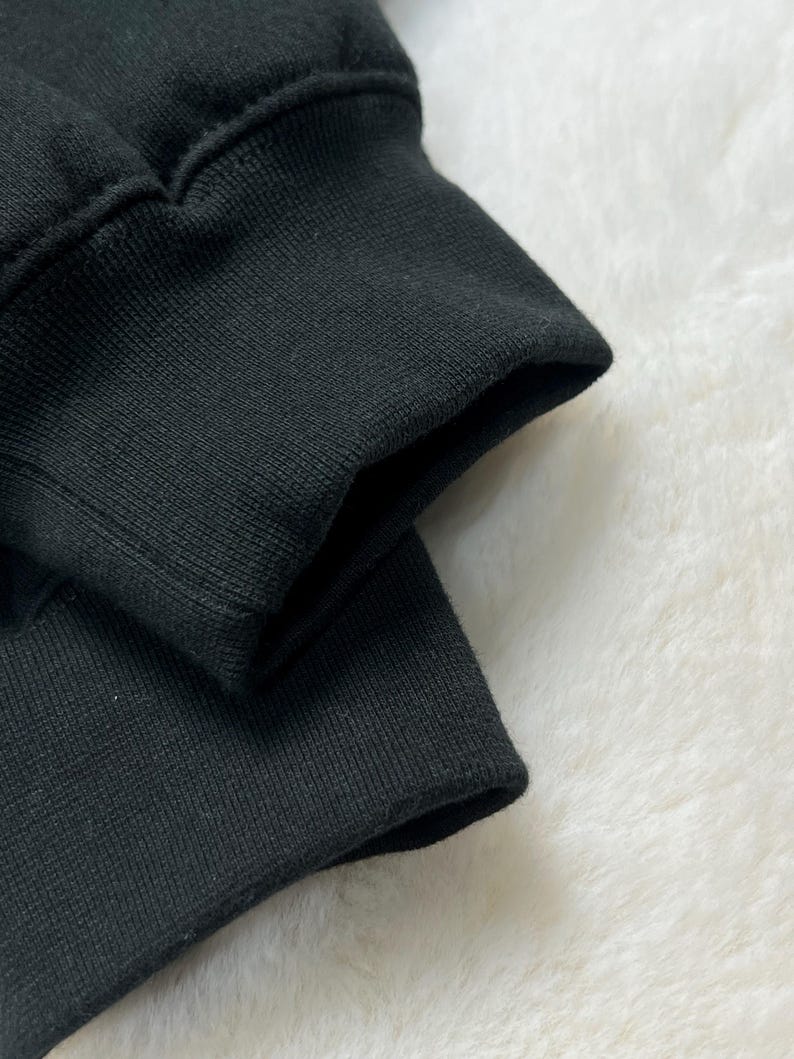 Puede incluir: Primer plano de un pu&ntilde;o de sudadera negro, que muestra la tela de punto acanalado y la costura donde se une a la manga. El pu&ntilde;o est&aacute; doblado, revelando el interior de la tela. El fondo es una superficie suave y blanca.
