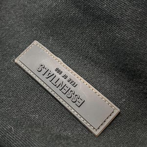 Puede incluir: Primer plano de una etiqueta rectangular gris con la palabra "ESSENTIALS" en relieve en gris oscuro. El texto "FEAR OF GOD" est&aacute; encima de la palabra "ESSENTIALS". La etiqueta est&aacute; cosida alrededor de los bordes y sobre un fondo de tela negra.