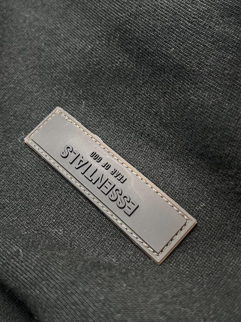 Puede incluir: Primer plano de una etiqueta rectangular gris con la palabra "ESSENTIALS" en relieve en gris oscuro. El texto "FEAR OF GOD" est&aacute; encima de la palabra "ESSENTIALS". La etiqueta est&aacute; cosida alrededor de los bordes y sobre un fondo de tela negra.