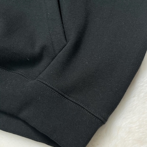 Puede incluir: Primer plano de una sudadera con capucha negra. La imagen resalta la textura de la tela y los detalles de las costuras. El pu&ntilde;o y el dobladillo acanalados son visibles, lo que indica un estilo informal.