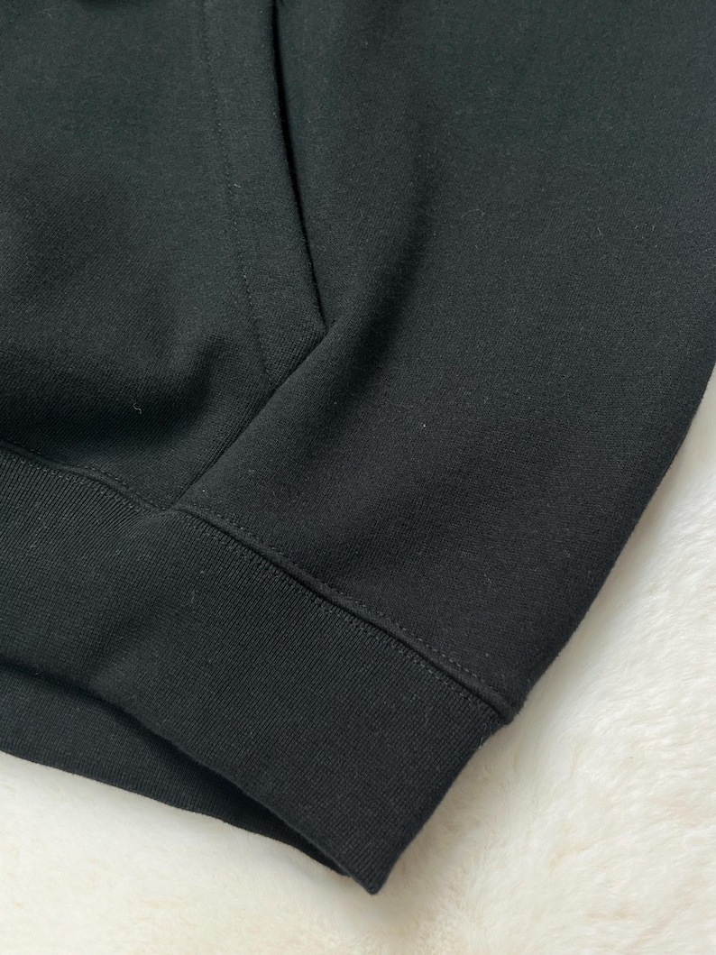 Puede incluir: Primer plano de una sudadera con capucha negra. La imagen resalta la textura de la tela y los detalles de las costuras. El pu&ntilde;o y el dobladillo acanalados son visibles, lo que indica un estilo informal.