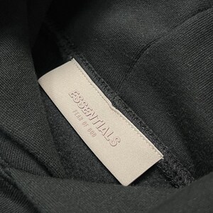 Puede incluir: Primer plano de una sudadera con capucha negra con una etiqueta rectangular de color beige claro. La etiqueta tiene la inscripci&oacute;n en relieve "ESSENTIALS FEAR OF GOD". La tela oscura de la sudadera crea un fuerte contraste.
