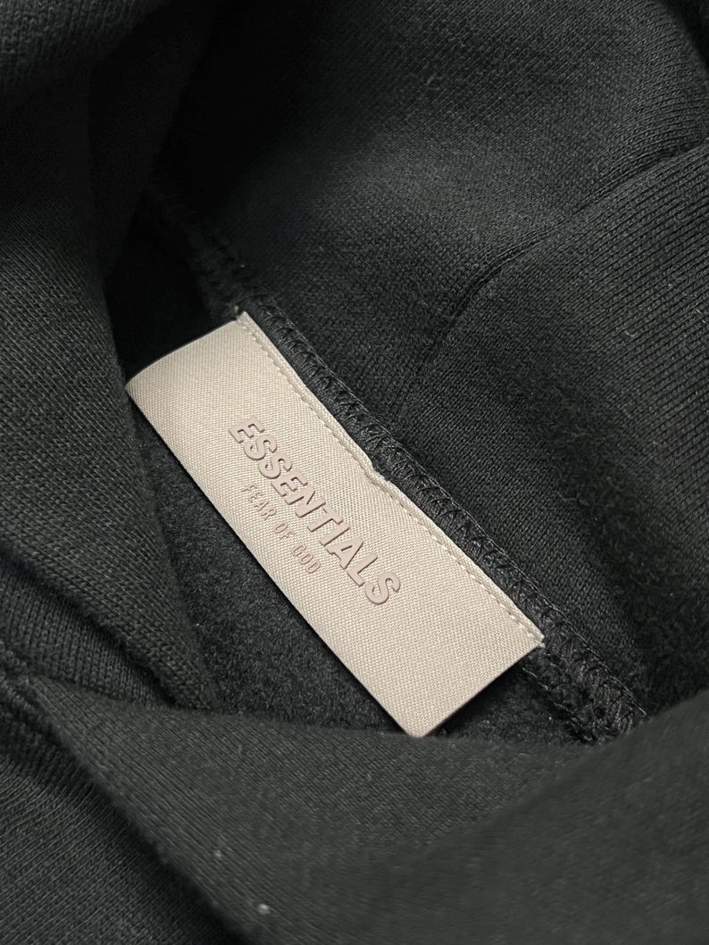 Puede incluir: Primer plano de una sudadera con capucha negra con una etiqueta rectangular de color beige claro. La etiqueta tiene la inscripci&oacute;n en relieve "ESSENTIALS FEAR OF GOD". La tela oscura de la sudadera crea un fuerte contraste.