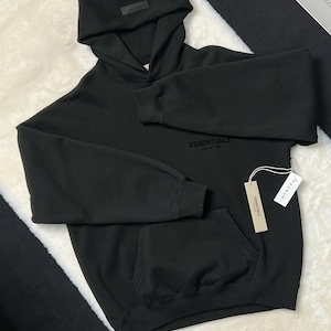 Puede incluir: Sudadera con capucha negra con bolsillo delantero y capucha adjunta. La palabra "ESSENTIALS" est&aacute; impresa en el pecho. Una etiqueta con la palabra "ESSENTIALS" est&aacute; adjunta a la sudadera con capucha.