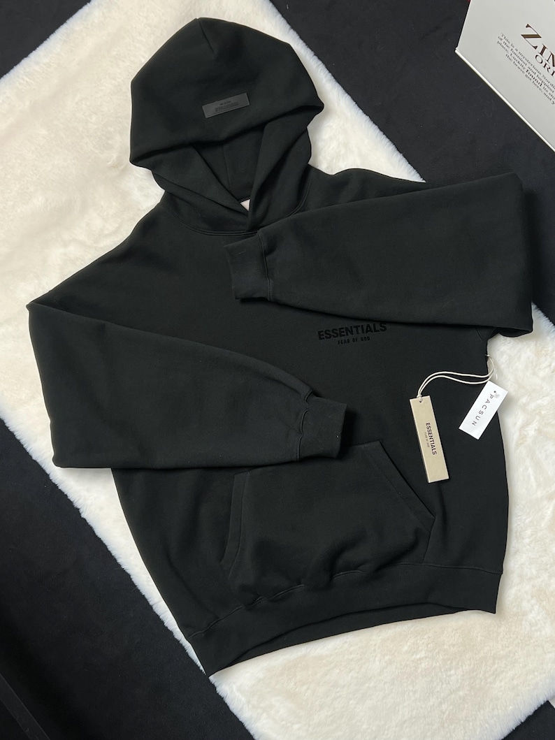 Puede incluir: Sudadera con capucha negra con bolsillo delantero y capucha adjunta. La palabra "ESSENTIALS" est&aacute; impresa en el pecho. Una etiqueta con la palabra "ESSENTIALS" est&aacute; adjunta a la sudadera con capucha.