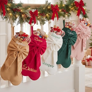 May include: Five velvet Christmas stockings in tan, red, white, dark green, and light pink, each featuring a large decorative bow and embroidered names: Sophia, Dorian, Jasper, Gideon, and Marius. The stockings are filled with assorted treats.