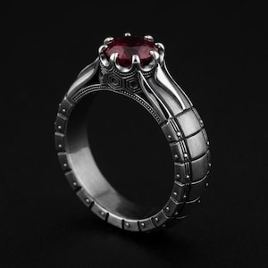 May include: A silver ring with a large, round, deep red gemstone. The ring has a detailed design with geometric patterns and a textured band. The setting has a crown-like appearance, and the ring is set against a black background.