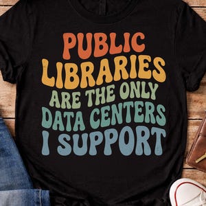 Könnte beinhalten: Ein schwarzes kurzärmeliges T-Shirt mit dem Text PUBLIC LIBRARIES ARE THE ONLY DATA CENTERS I SUPPORT in einer gestapelten, wellenförmigen Schriftart. Die Buchstaben sind in Orange, Gelb, Hellgrün und Hellblau auf dem schwarzen Stoff gehalten.