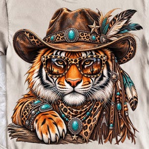 Könnte beinhalten: Ein beiges T-Shirt mit einer Grafik eines Tigers, der einen braunen Cowboyhut mit Leopardenmuster, Federn, türkisfarbenen Akzenten und einem Stern trägt. Der Tiger trägt eine Sonnenbrille mit Leopardenmuster, die eine Wüstenlandschaft reflektiert.
