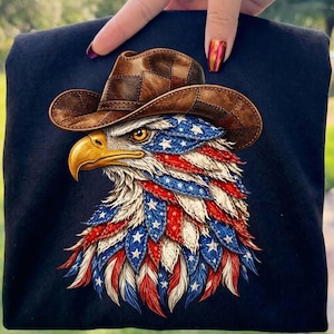Op de afbeelding: Marineblauw T-shirt met een gedetailleerde afbeelding van een adelaar met een cowboyhoed. De veren van de adelaar zijn ontworpen met de kleuren en sterren van de Amerikaanse vlag. De adelaar heeft een gele snavel en een bruine cowboyhoed.