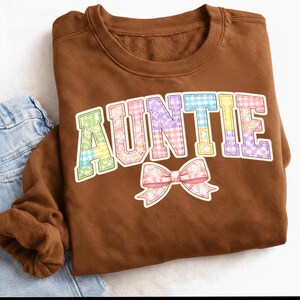 May include: A brown sweatshirt with the word "AUNTIE" in colorful patchwork letters and a matching bow graphic. The sweatshirt is folded, with a portion of blue denim jeans visible in the corner.