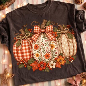 May include: A dark grey short-sleeved t-shirt featuring a graphic print of three pumpkins. The left pumpkin has an orange gingham pattern, the centre has a floral design with a red and white plaid bow, and the right has a beige polka dot pattern.