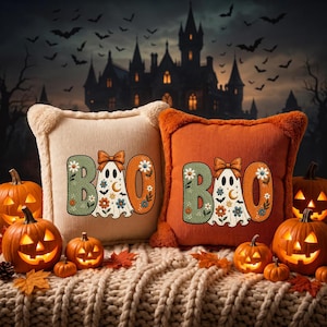 May include: Two square throw pillows, one cream and one orange, featuring embroidered text that reads BOO with a ghost wearing a bow in the center. The ghost is decorated with floral patterns, moons, and bats. The pillows have fuzzy edges.