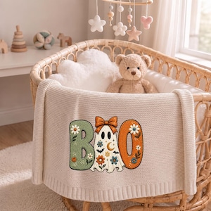 May include: A cream-colored knit baby blanket featuring an embroidered design of the word BOO. The letter B is sage green with floral accents, the letter O is orange with floral accents, and a ghost with a bow and stars replaces the middle O.